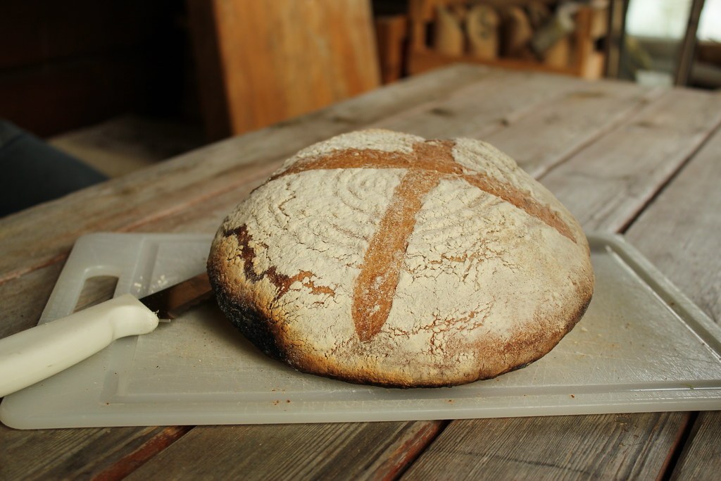A round sourdough loaf scored in a cross, dusted with flour, on a board on a plank table
