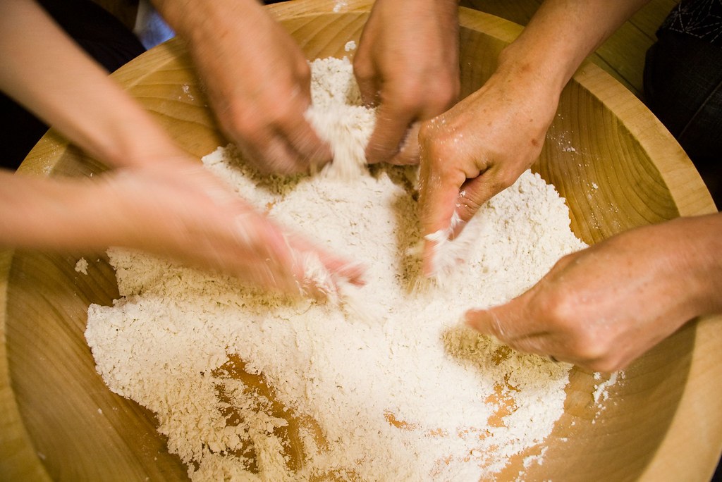 Four pairs of hands working flour together in a wide wooden bowl