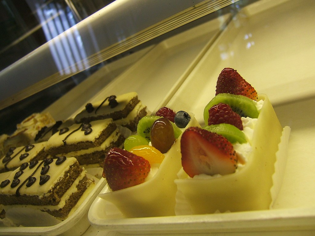 Fruit tarts topped with strawberry and kiwi beside iced cake slices in a counter tray